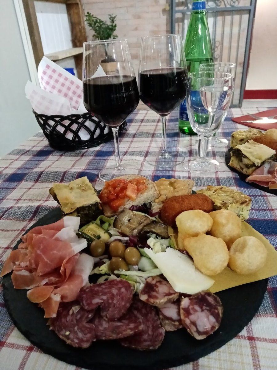 Il tagliere dell'antipasto della casa: prosciutto crudo, salame, olive, formaggio, verdure grigliate, frittata e pizza fritta, con due calici di vino rosso.
