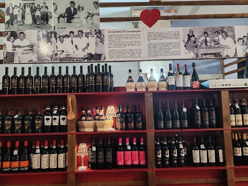 La parete della cantina: scaffali rossi pieni di bottiglie e, sopra, un pannello con le fotografie di famiglia in bianco e nero.